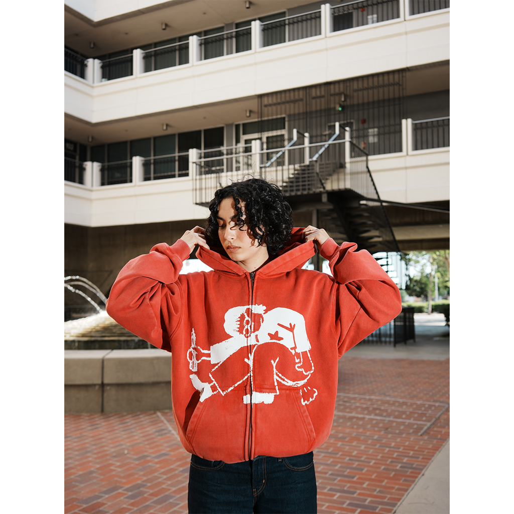 Model wearing a red hoodie with a white graphic design in an outdoor setting with hands in pocket.