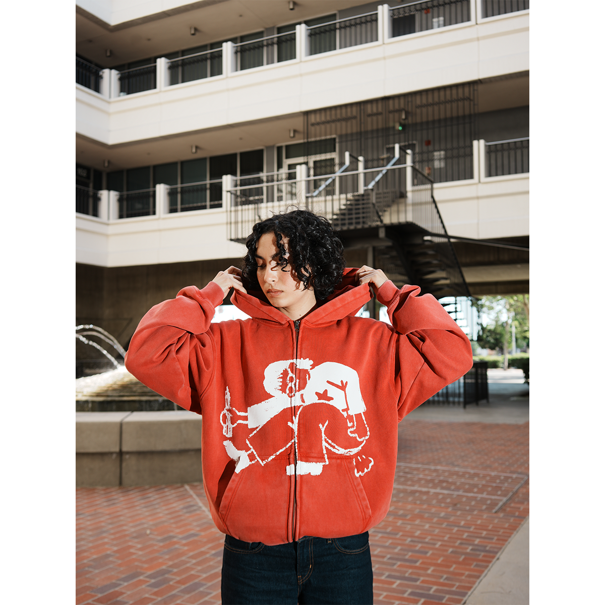 Model wearing a red hoodie with a white graphic design in an outdoor setting with hands in pocket.