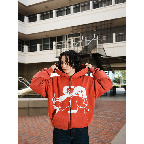 Model wearing a red hoodie with a white graphic design in an outdoor setting with hands in pocket.